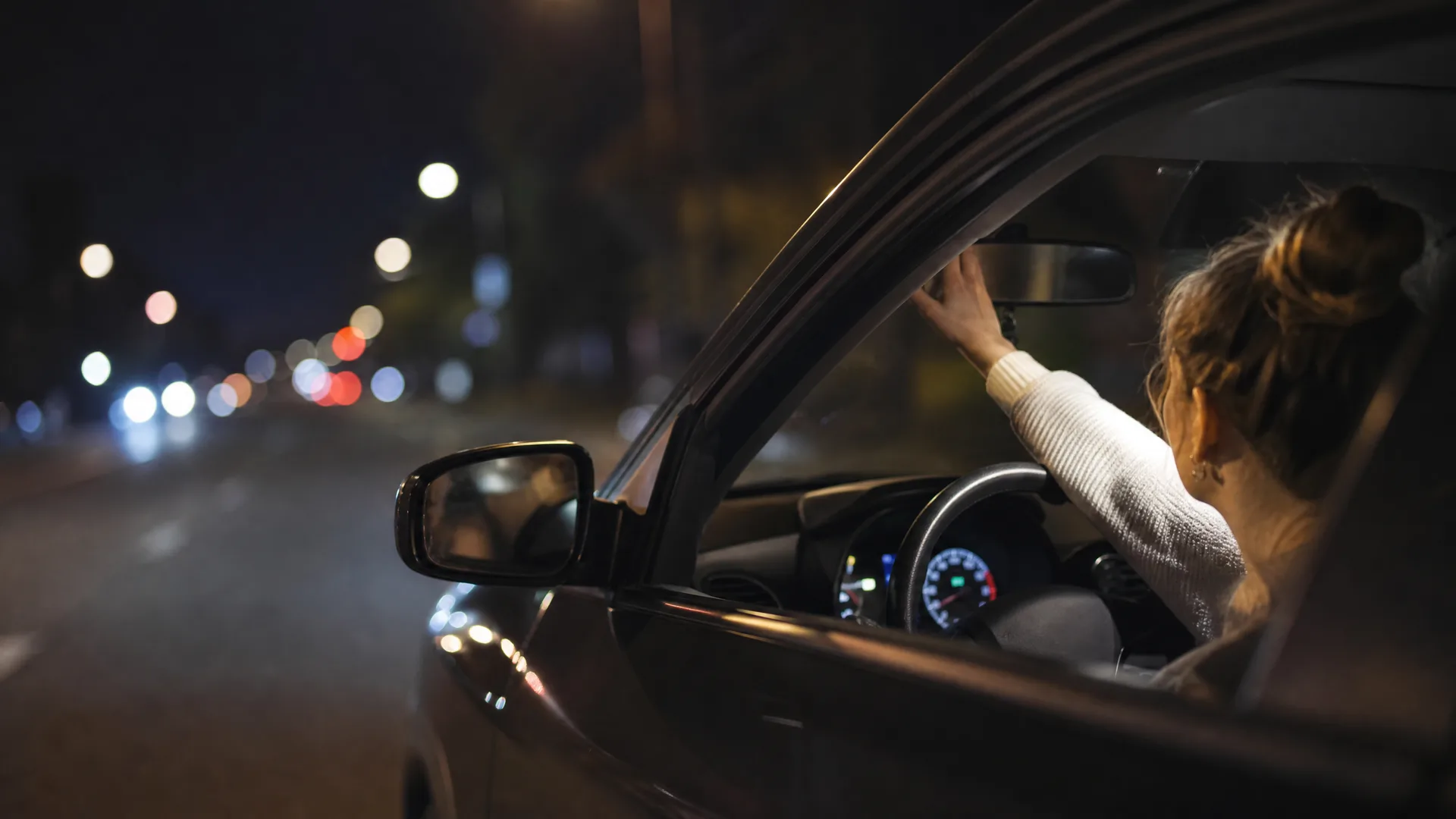 A woman in a white sweater sits in a car's driver seat at night, one hand on the steering wheel and the other raised toward the rearview mirror, with blurred street lights visible through the open window.