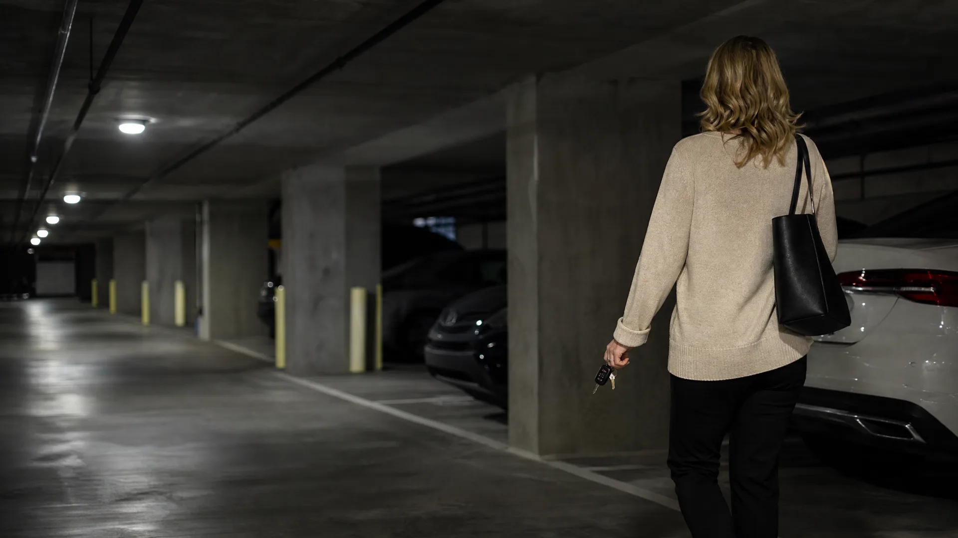A woman in a beige sweater and black pants walks away from the camera through a dimly lit concrete parking garage, carrying a black tote bag and holding car keys.