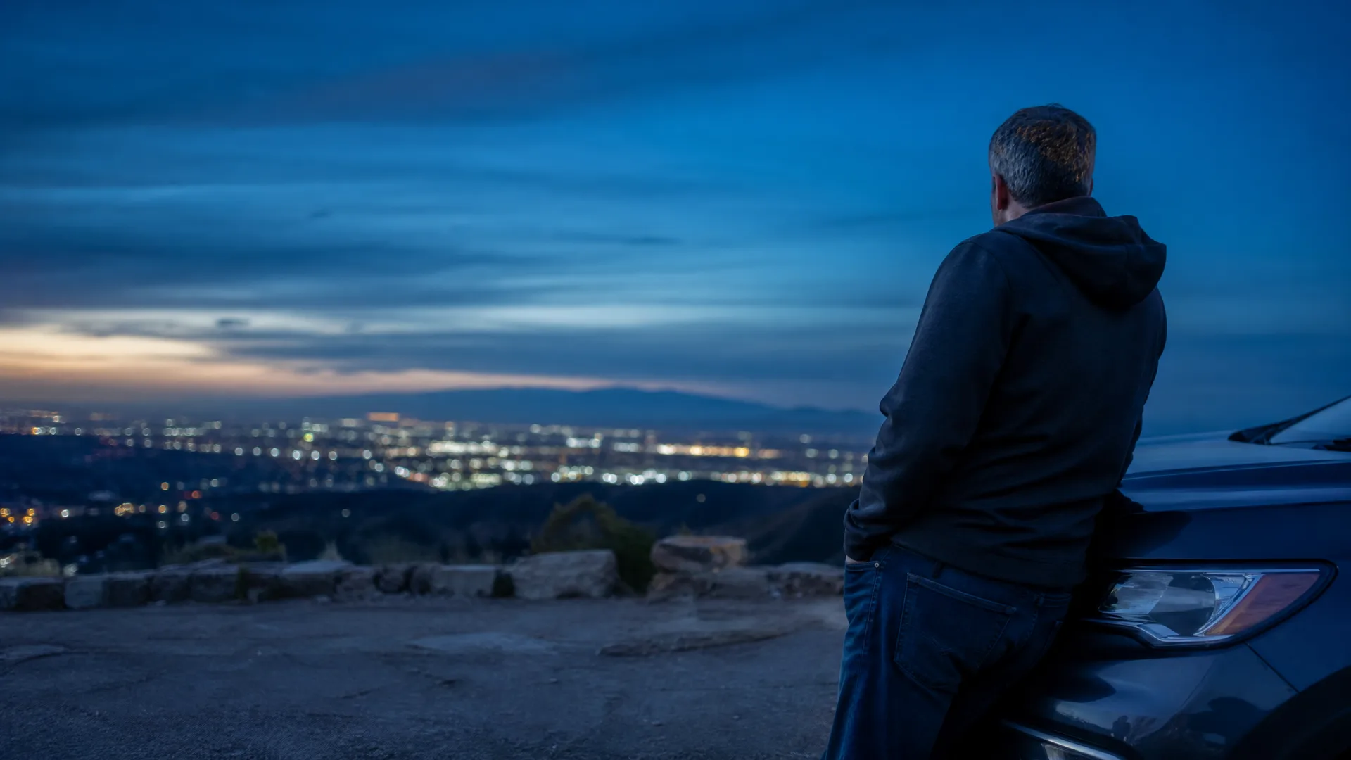 A man in a dark hooded jacket leans against a car at a hilltop overlook at dusk, facing away toward distant city lights under a deep blue sky.