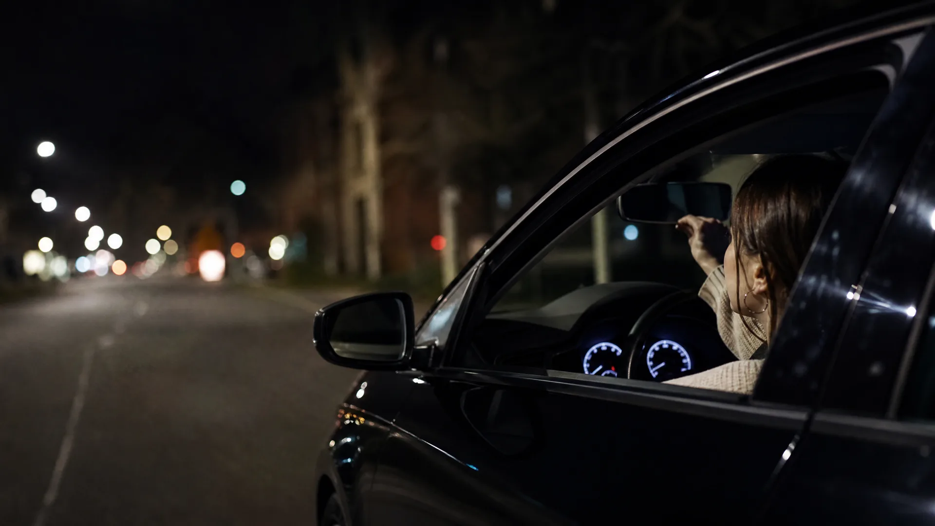 At night, a woman sits in the driver's seat of a dark car with the window open, reaching toward the rearview mirror, with lit instrument dials visible and blurred street lights along the road behind.