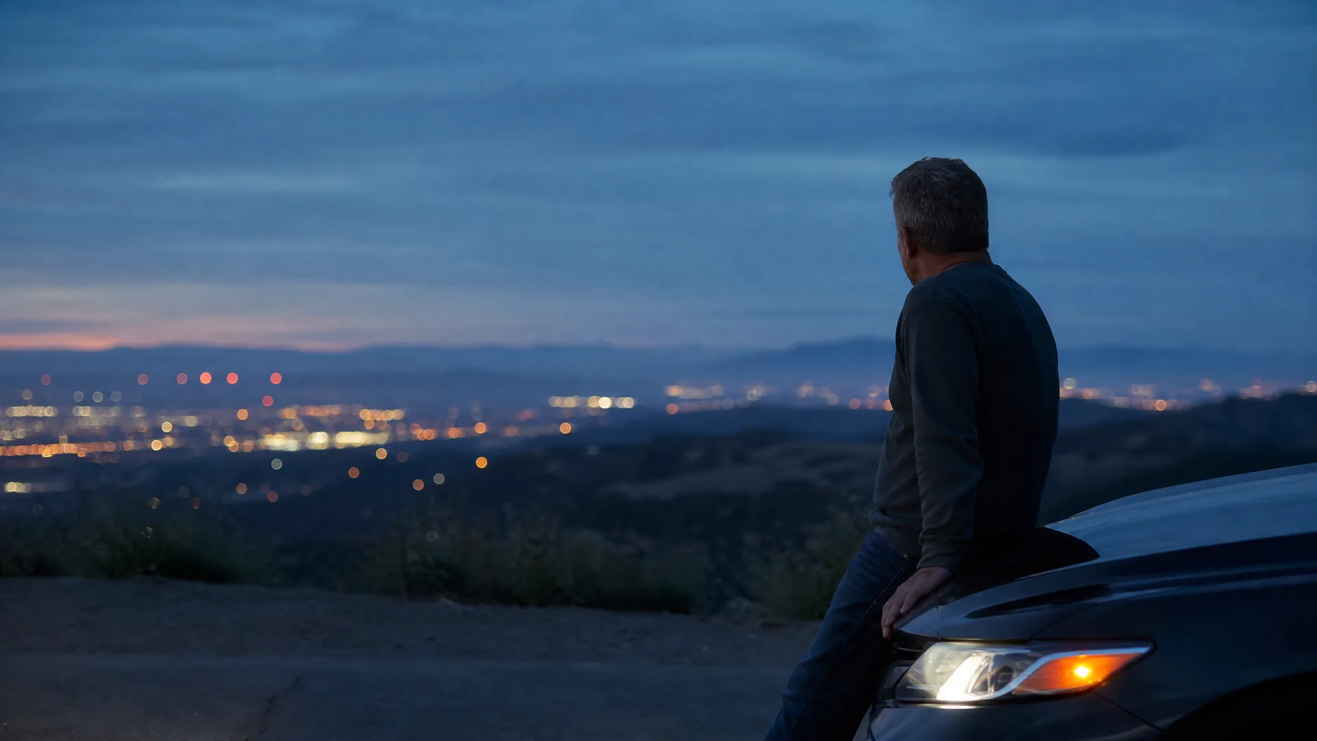 A man in a dark long-sleeved shirt and jeans leans against the front of a car on a hilltop at dusk, looking out over distant city lights.