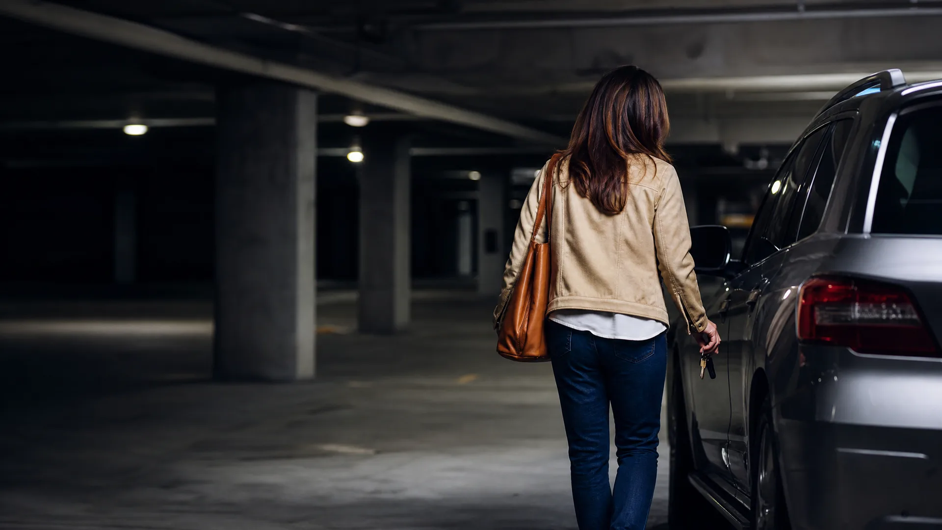 A woman seen from behind in a beige jacket, jeans and tan shoulder bag walks toward a parked car holding keys in a dimly lit underground parking garage with concrete columns.