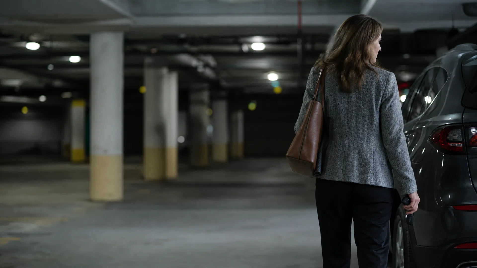 A woman in a grey blazer and black trousers with a brown shoulder bag stands beside a parked car in an underground parking garage, looking back over her shoulder.
