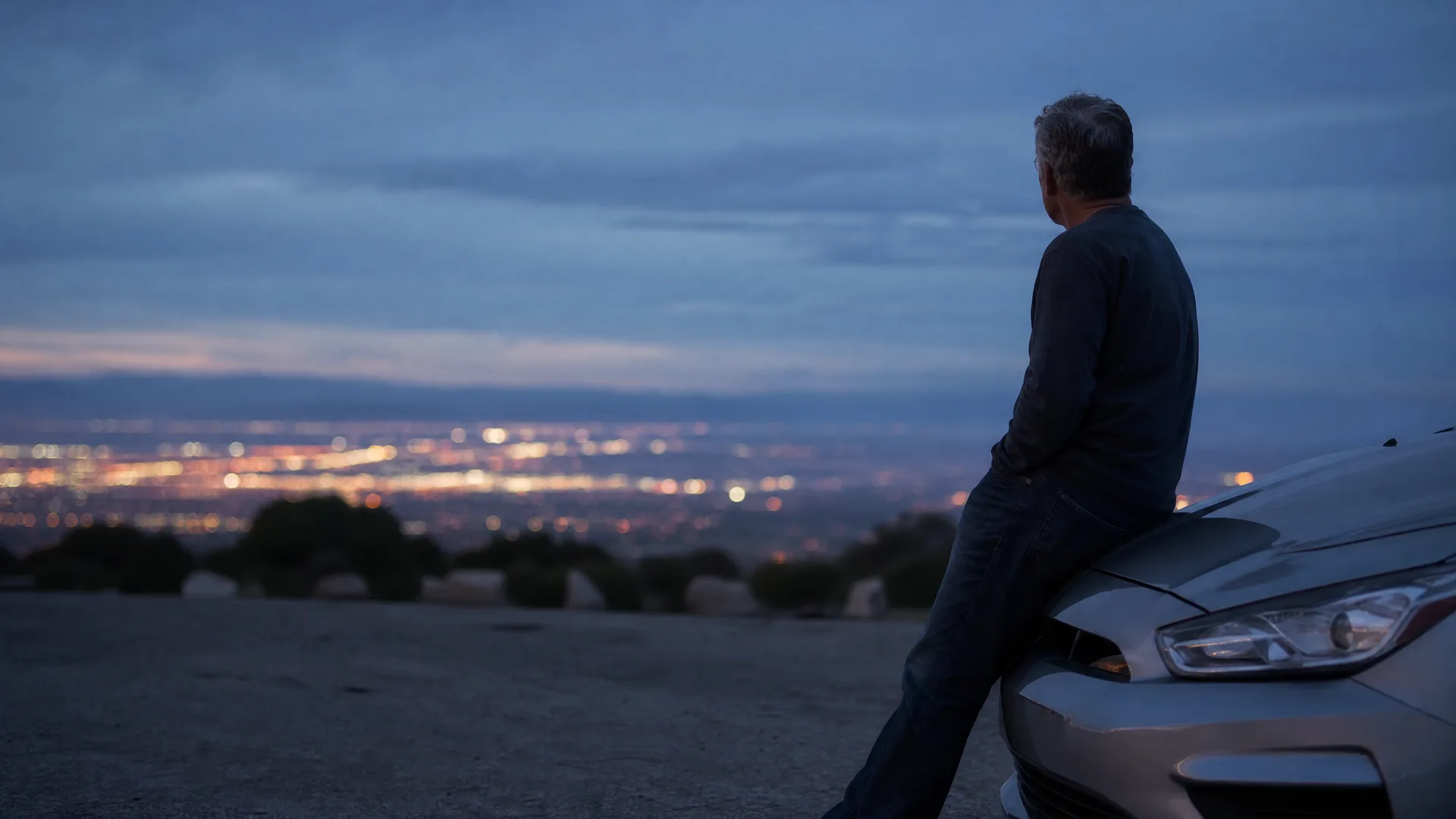 A man in a dark sweater leans against the front of a silver car on a gravel overlook at dusk, facing distant city lights below.