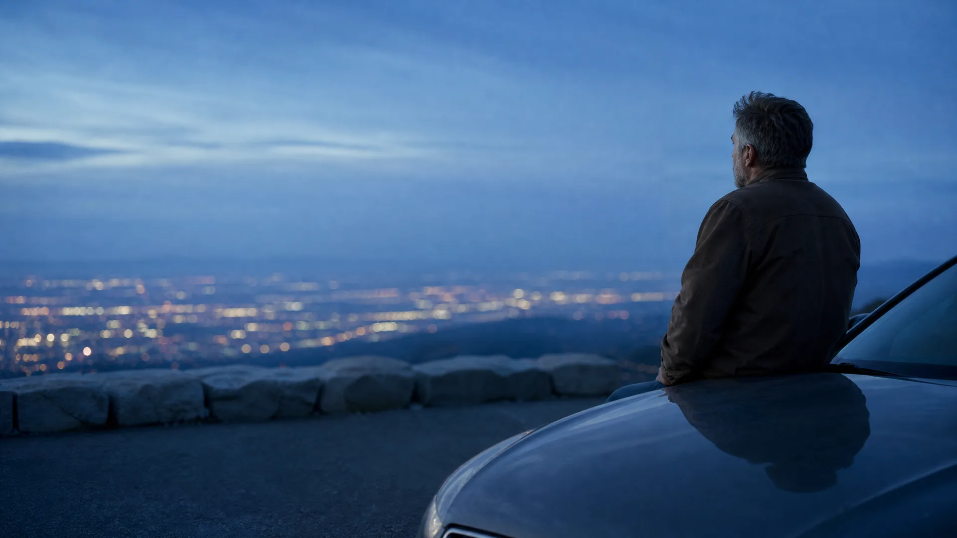 A man in a dark jacket sits on the hood of a car at a hilltop overlook at dusk, looking out over city lights below a blue sky.