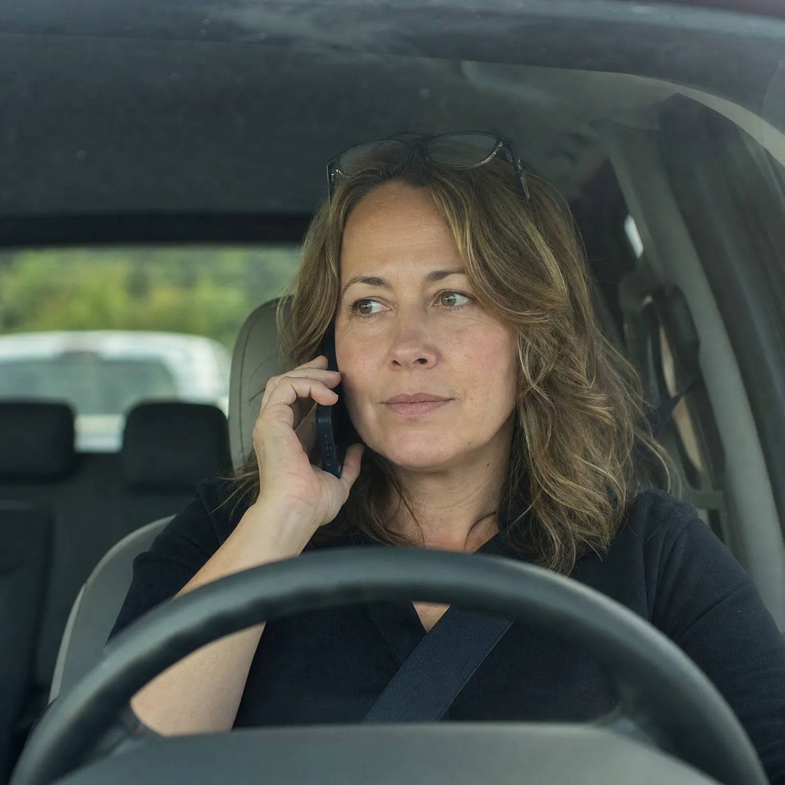 A woman with wavy brown hair and sunglasses pushed onto her head sits in a car's driver seat, holding a phone to her ear with one hand while looking out the side window.