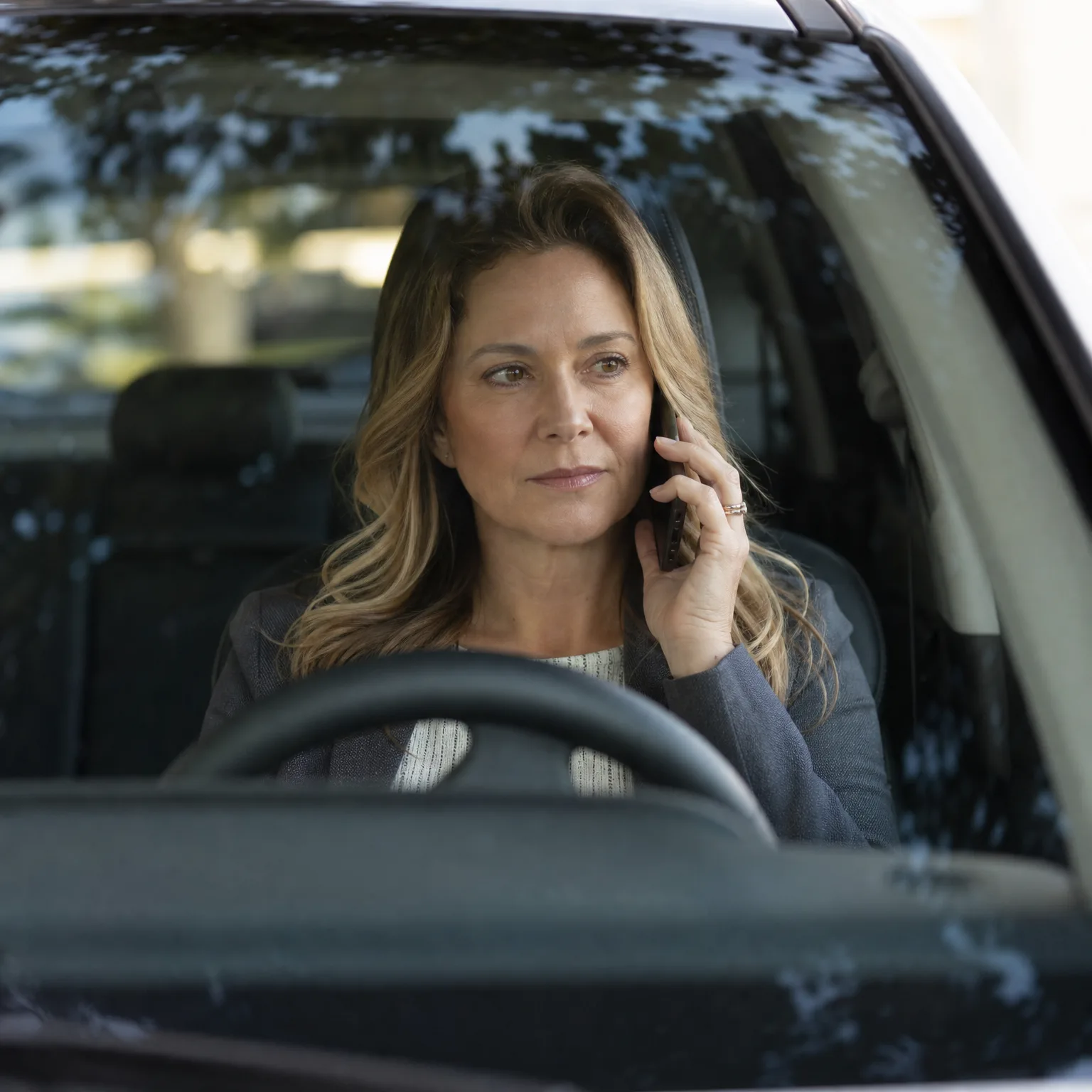 A woman with long blonde-brown hair sits in a car's driver seat behind the steering wheel, holding a phone to her ear and looking to the side, seen through the windshield.