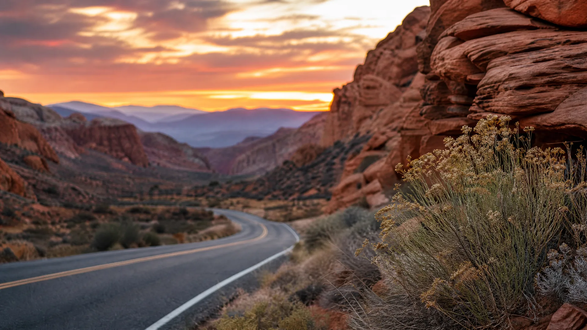 A two-lane paved road curves through a red rock canyon with desert scrub in the foreground and distant layered mountains under an orange sunset sky.