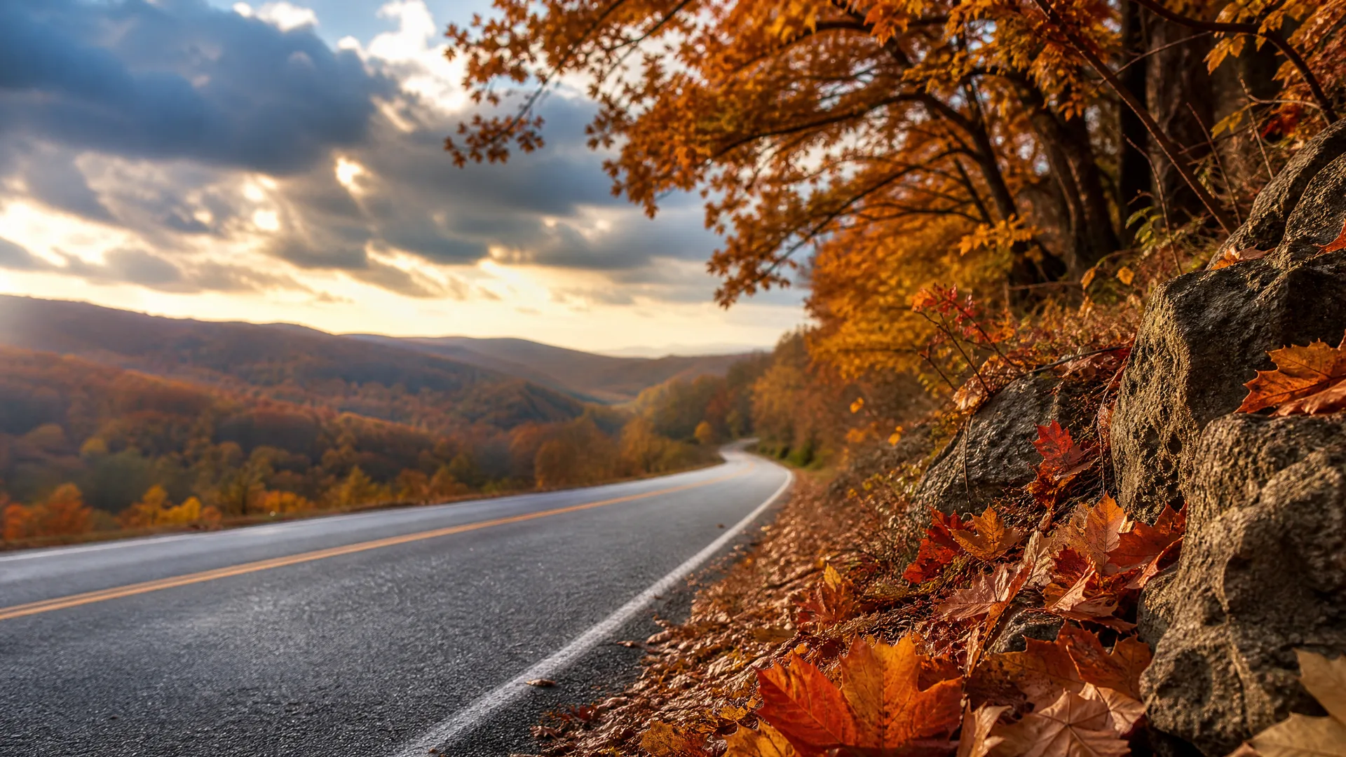 A curving asphalt road with yellow center lines and a white edge line runs past a rocky bank covered in fallen orange leaves, with autumn-colored trees overhead and forested hills under a cloudy sky at low sun.