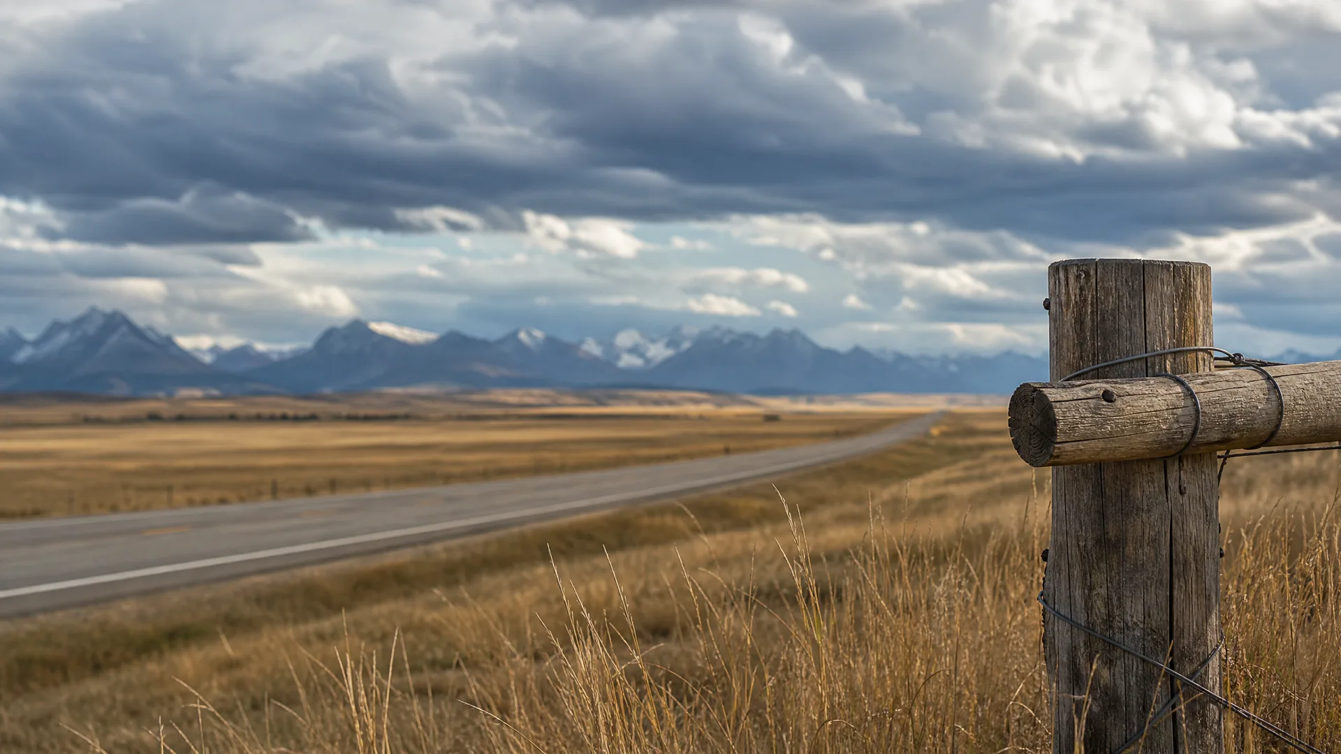 A weathered wooden fence post with wire in the right foreground beside a two-lane road crossing dry golden grassland, with a snow-dusted mountain range and heavy clouds in the distance.