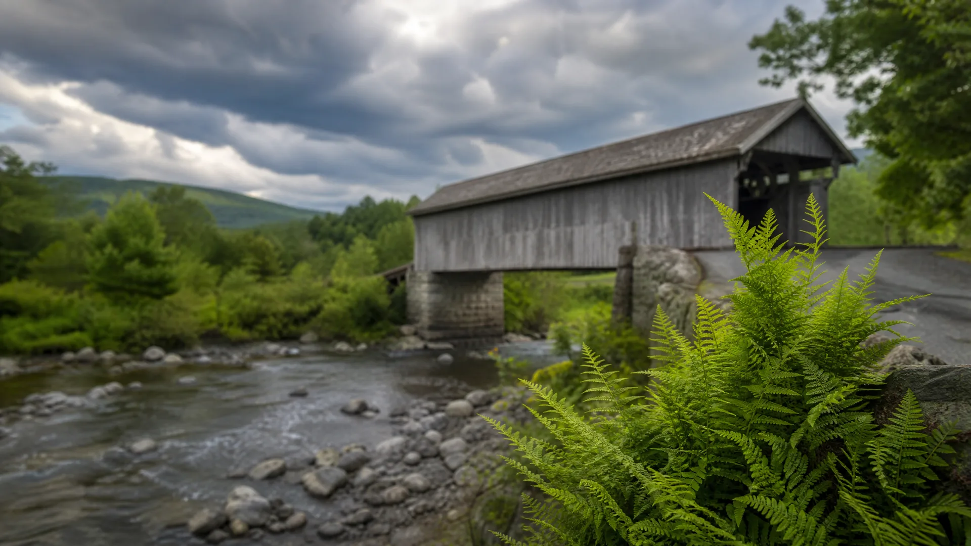A weathered wooden covered bridge on stone piers spans a rocky shallow river under a cloudy sky, with green ferns in the foreground and forested hills behind.