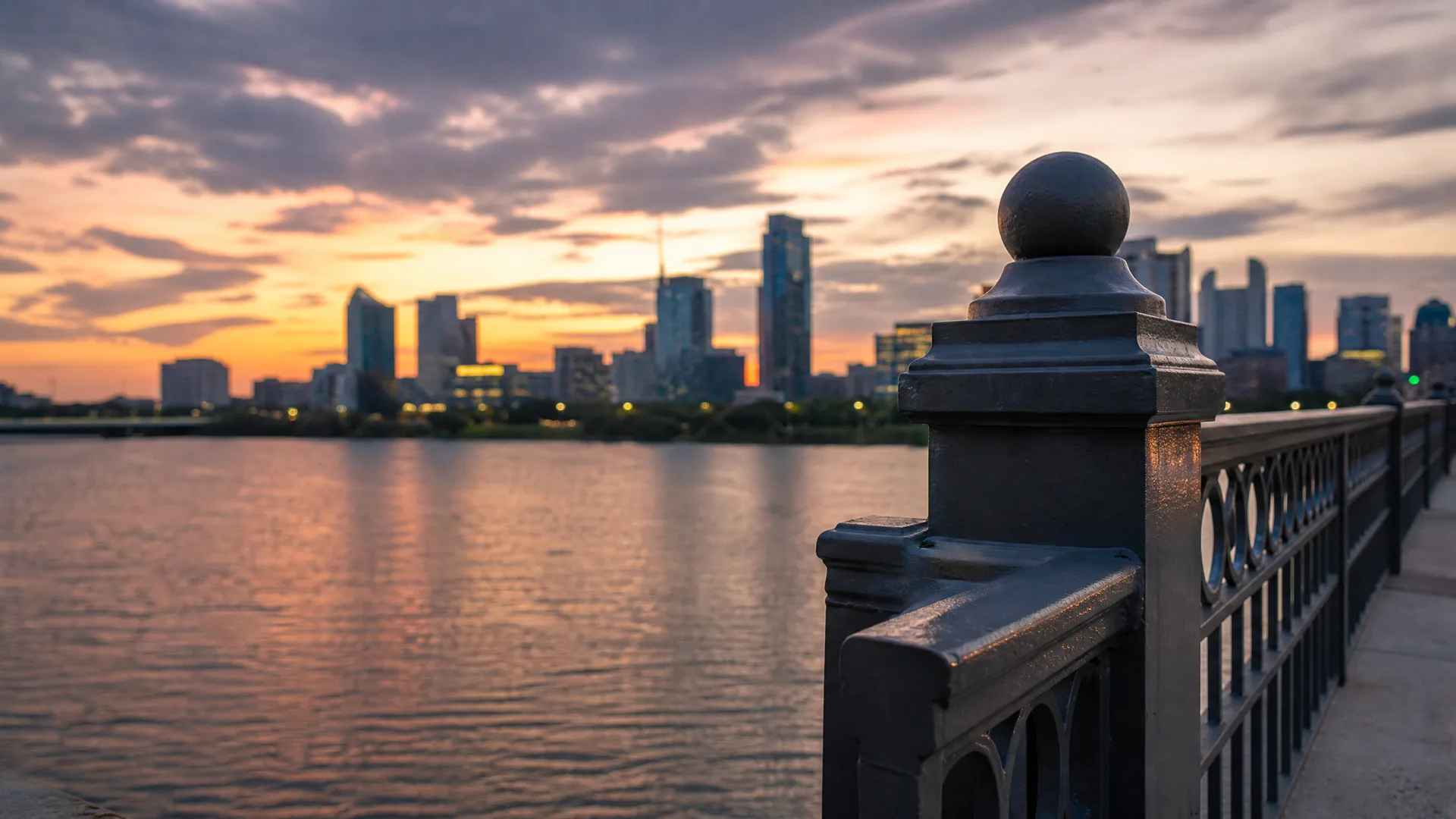 A dark metal railing post with a ball finial in sharp foreground focus along a waterfront walkway, with a blurred city skyline across calm water under an orange and purple sunset sky.
